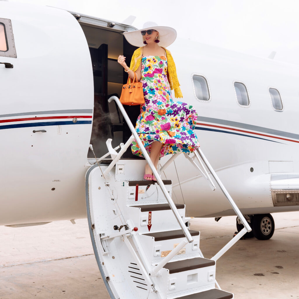 Sheree Frede of the SheShe Show standing on stairs leading into private jet wearing a floral dress and big white hat!