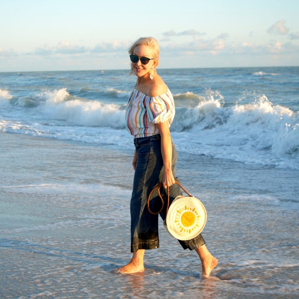 stripe off the shoulder top, cropped denim jeans with button fly, straw circle bag, barefoot walking along the beach