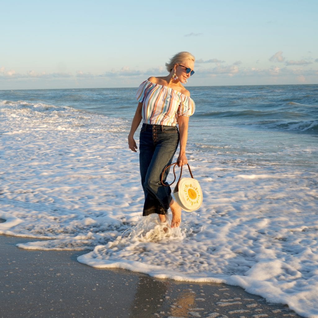 stripe off the shoulder top, cropped denim jeans with button fly, straw circle bag, barefoot walking along the beach