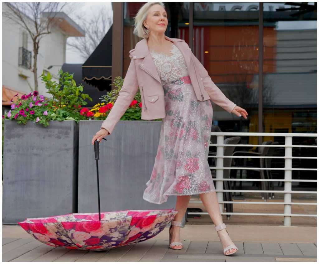 Sheree Frede standing in front of pretty spring flowers wearing a pink floral skirt and blush leather jacket and pink floral umbrella