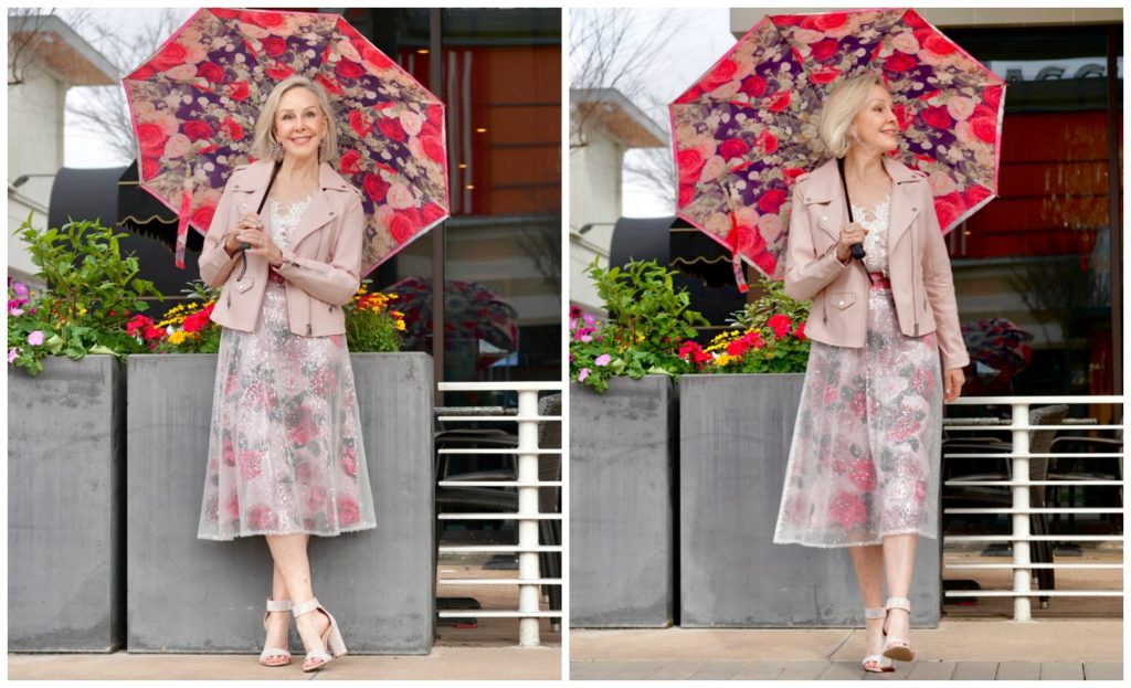 Sheree Frede standing in front of pretty spring flowers wearing a pink floral skirt and blush leather jacket and pink floral umbrella