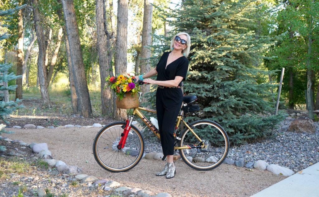 Sheree of the SheShe Show riding a bike with flowers in basket wearing a black jumpsuit
