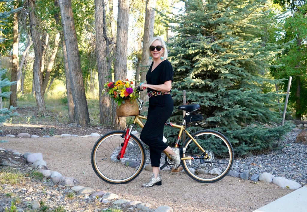 Sheree of the SheShe Show riding a bike with flowers in basket wearing a black jumpsuit