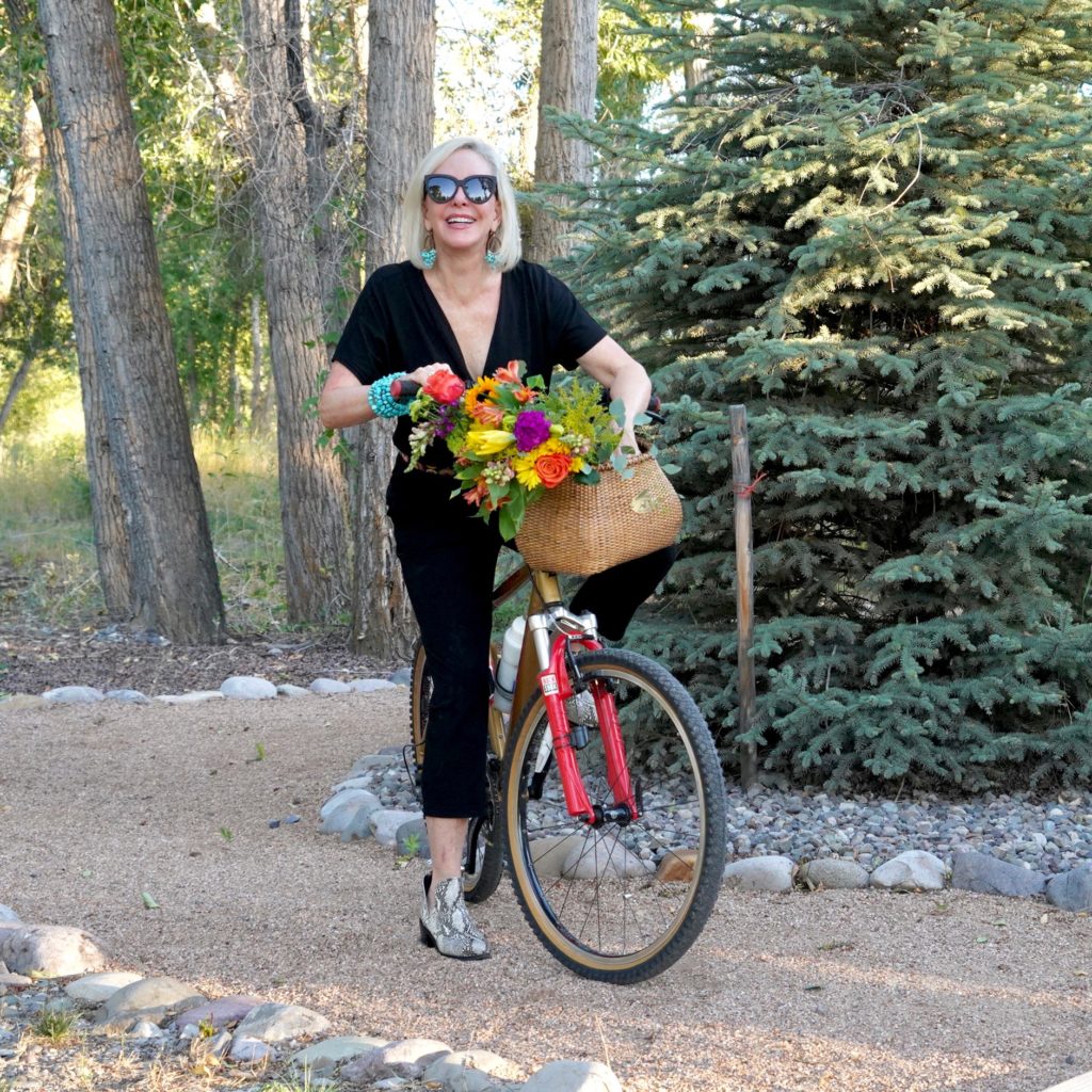 Sheree of the SheShe Show riding a bike with flowers in basket wearing a black jumpsuit