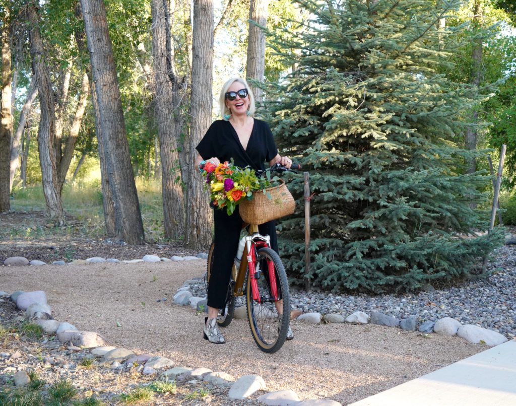 Sheree of the SheShe Show riding a bike with flowers in basket wearing a black jumpsuit