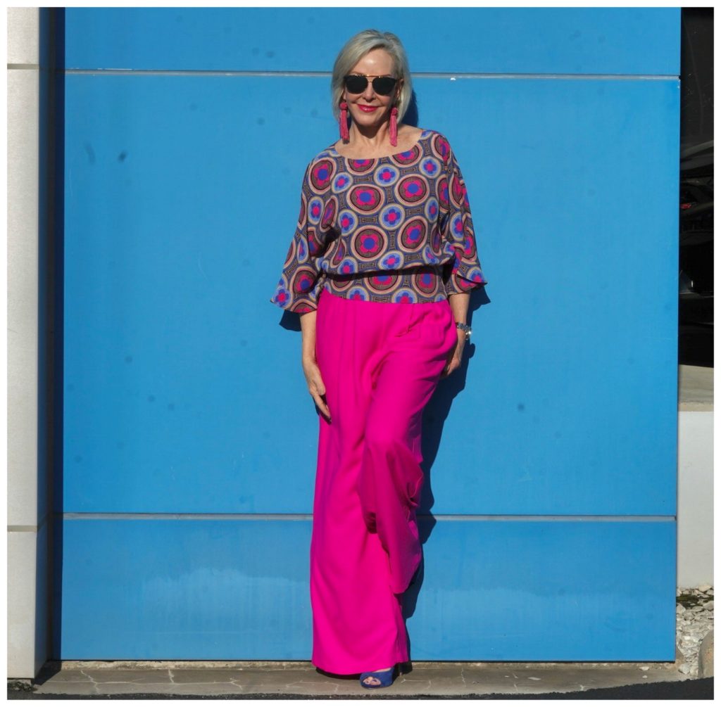 Sheree of the SheShe Show wearing hot pink wide leg pants and blue print top standing in front of blue wall