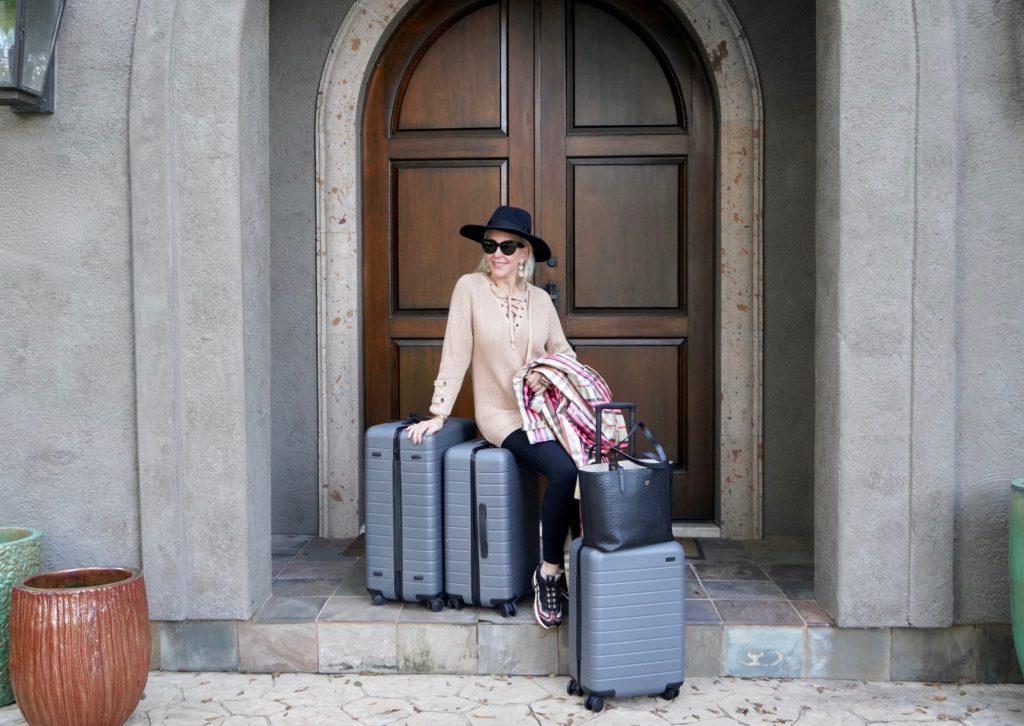 Sheree of the SheShe Show sitting with 3 large suitcases at her front door wearing black leggings, black hat and tan sweater.