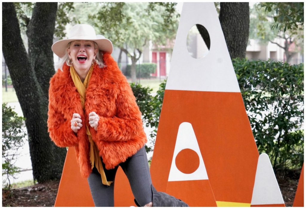 Sheree Frede of the SheShe Show standing in a candy corn patch wearing an orange faux fur jacket, jeans and white hat.