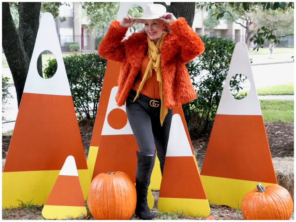 Sheree Frede of the SheShe Show standing in a candy corn patch wearing an orange faux fur jacket, jeans and white hat.