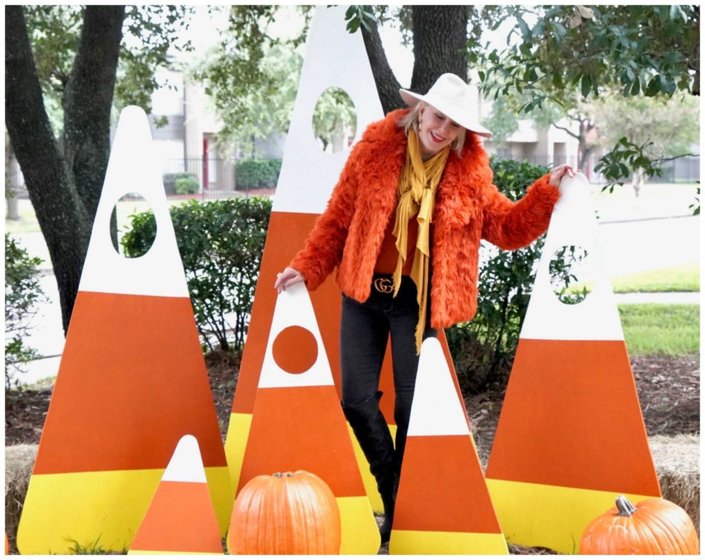 Sheree Frede of the SheShe Show standing in a candy corn patch wearing an orange faux fur jacket, jeans and white hat.