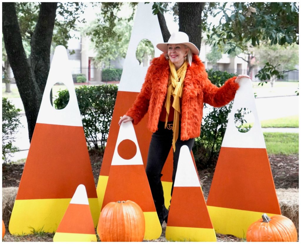 Sheree Frede of the SheShe Show standing in a candy corn patch wearing an orange faux fur jacket, jeans and white hat.