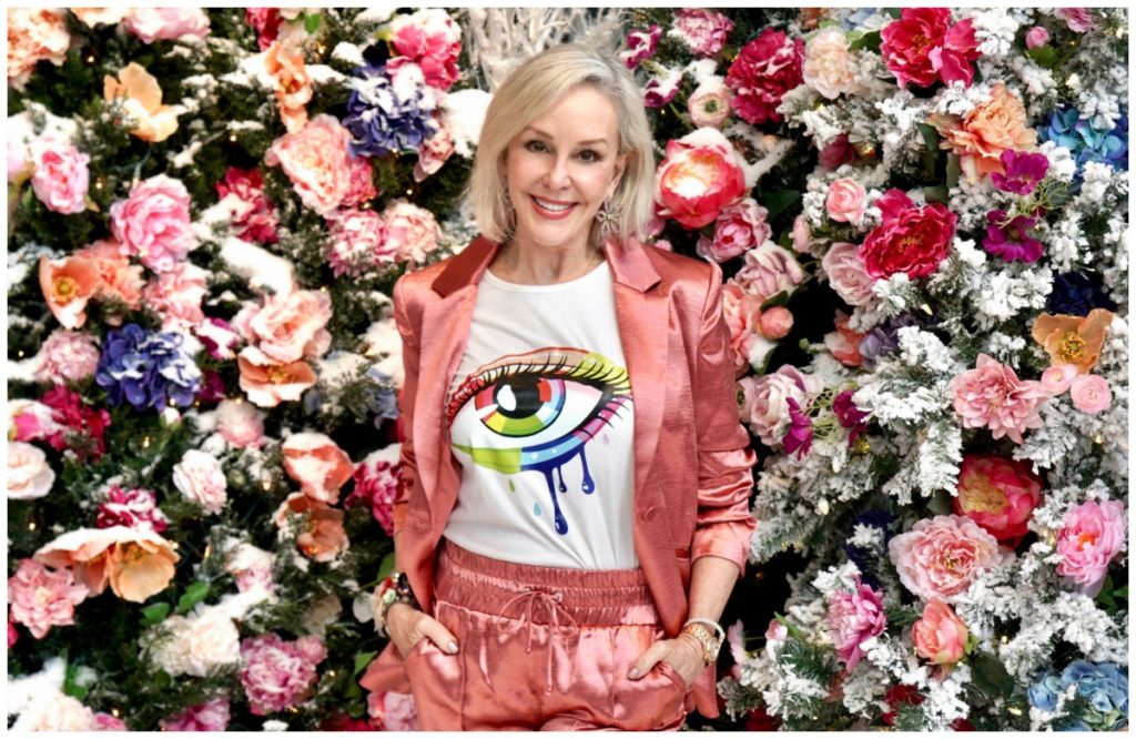 Sheree Frede of the SheSheShow weqring a satin 2 pc pant suit standing in a room of flowers and 2 stuffed polar bear animals