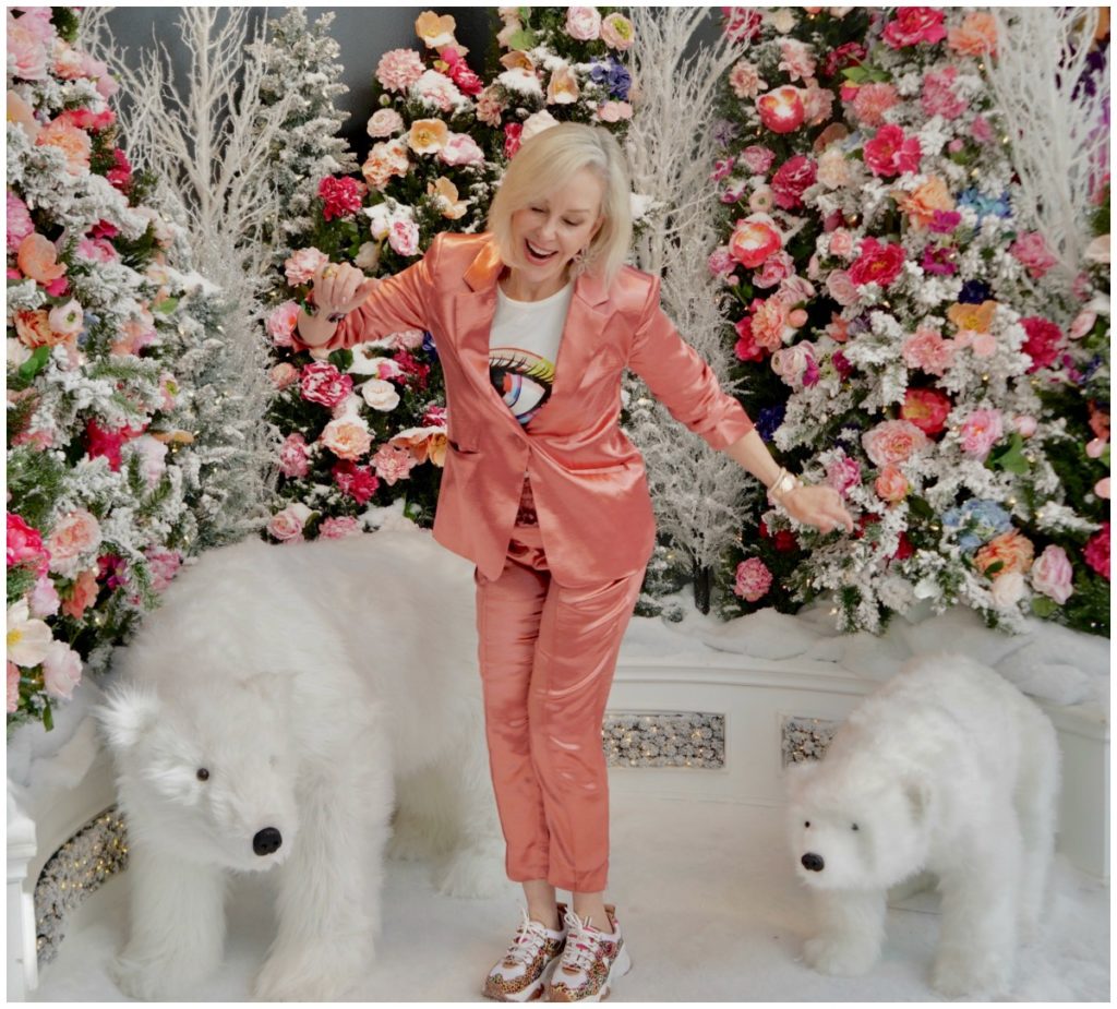 Sheree Frede of the SheSheShow weqring a satin 2 pc pant suit standing in a room of flowers and 2 stuffed polar bear animals