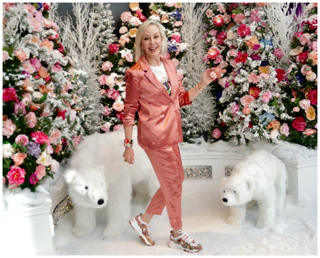 Sheree Frede of the SheSheShow weqring a satin 2 pc pant suit standing in a room of flowers and 2 stuffed polar bear animals