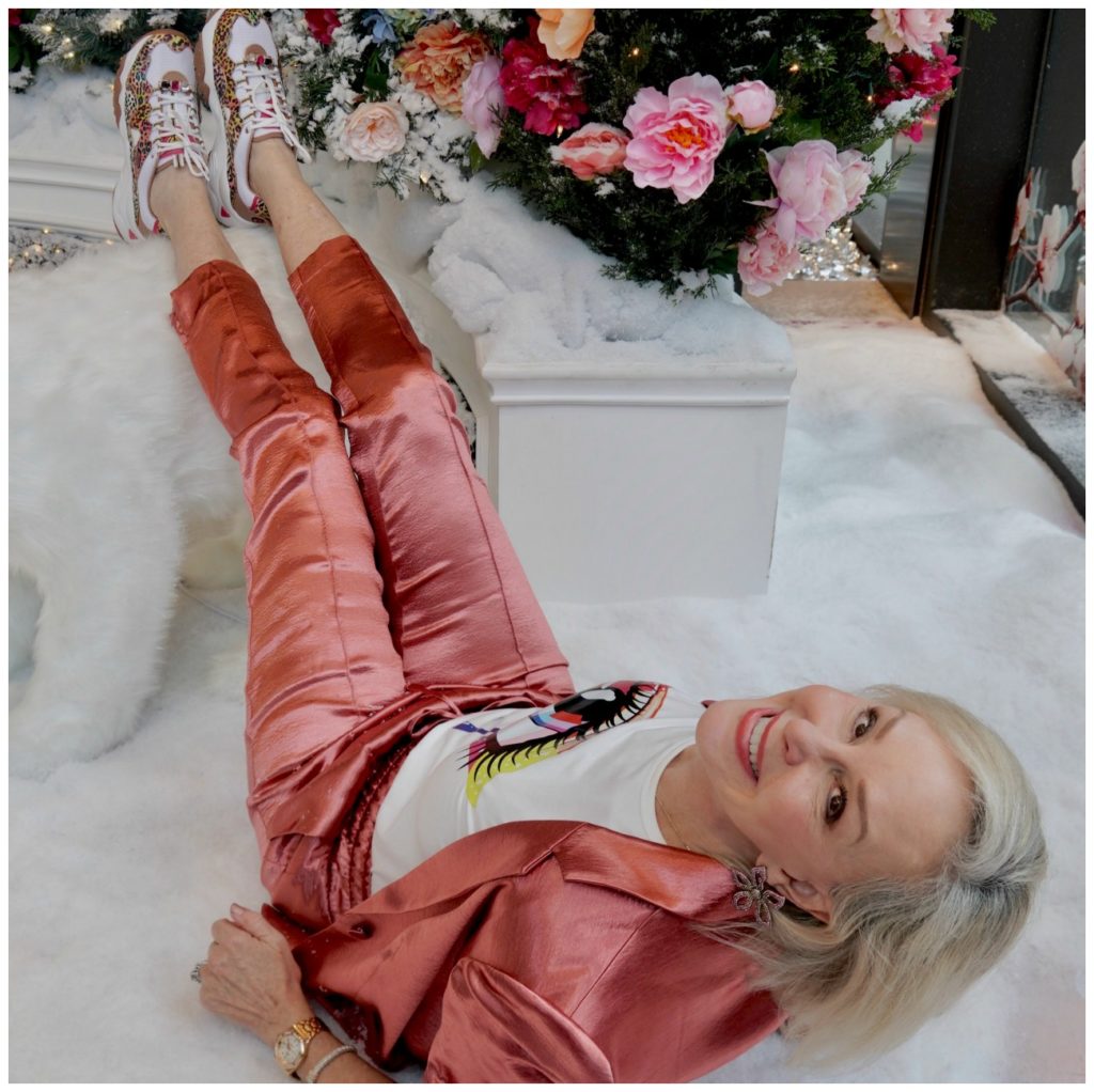 Sheree Frede of the SheSheShow weqring a satin 2 pc pant suit standing in a room of flowers and 2 stuffed polar bear animals