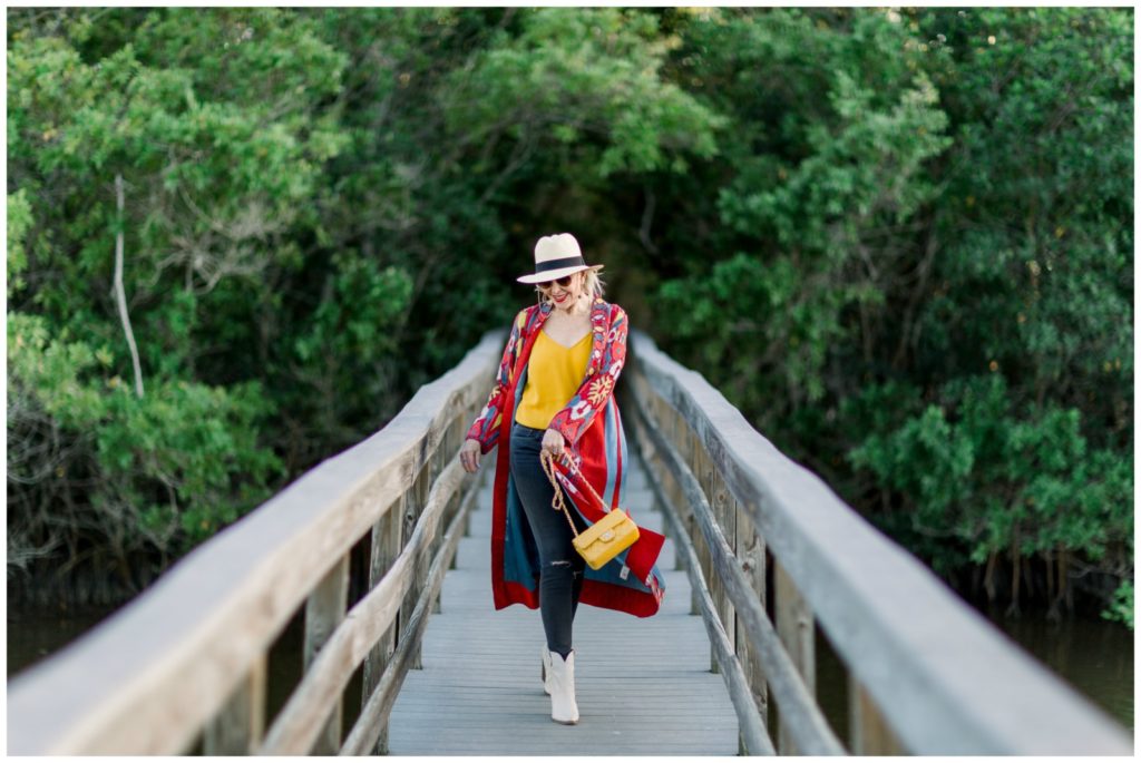 Sheree of the SheSheShow walking on wood walking bridge wearing a red print duster over  blue jeans and yellow camisole. Panama hat