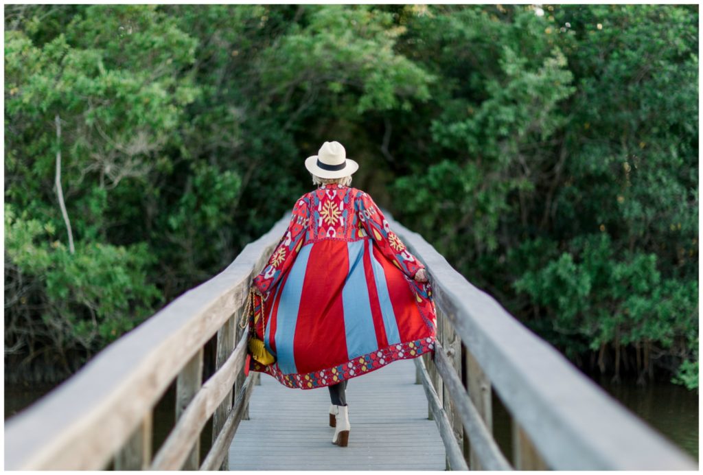 Sheree of the SheSheShow walking on wood walking bridge wearing a red print duster over blue jeans and yellow camisole. Panama hat