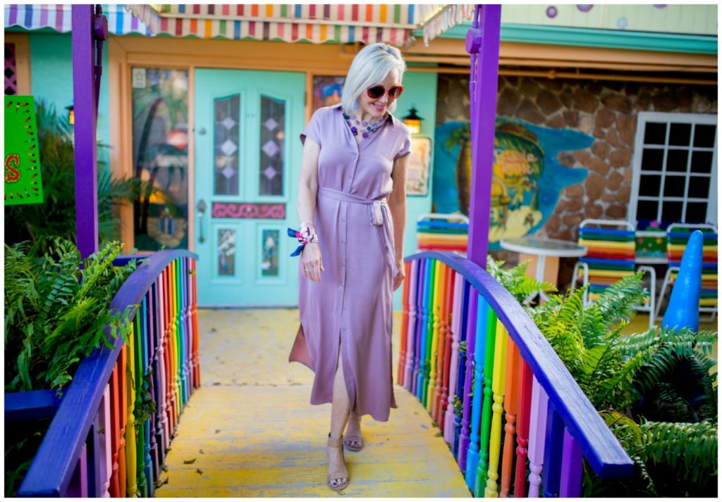 Sheree Frede of the SheSheShow walking across a bridge with rainbow railing wearing a lilac dress by Chicos