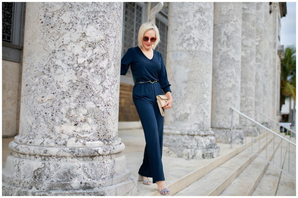 Sheree Frede of the SheSheShow standing by large stone columns and steps wearing asimple navy jumpsuit