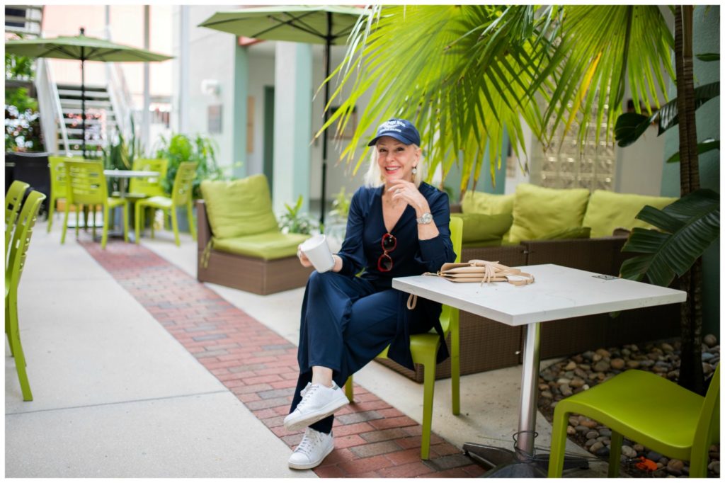 Sheree Frede of the SheSheShow in outdoor courtyeard wearing a simple navy jumpsuit and baseball cap