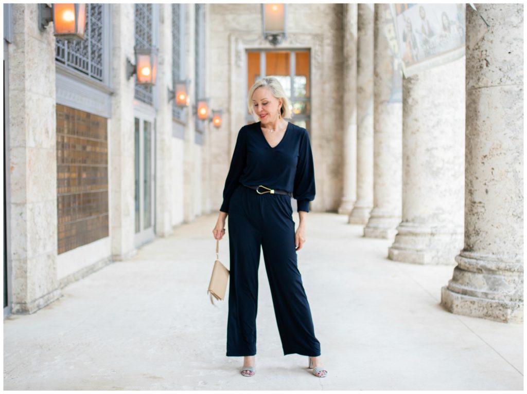 Sheree Frede of the SheSheShow standing by large stone columns wearing a simple navy jumpsuit