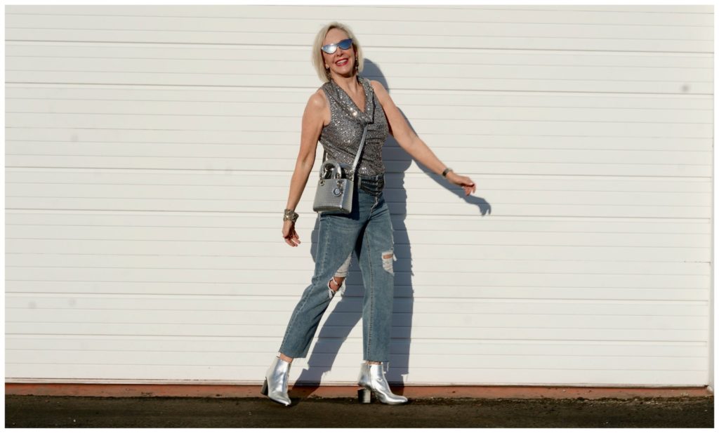 Sheree Frede of the SheShe Show standing in front of a white wall wearing gray sequin cowl neck top by Eliza J with ripped denim jean and silver booties