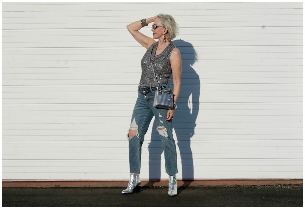 Sheree Frede of the SheShe Show standing in front of a white wall wearing gray sequin cowl neck top by Eliza J with ripped denim jean and silver booties