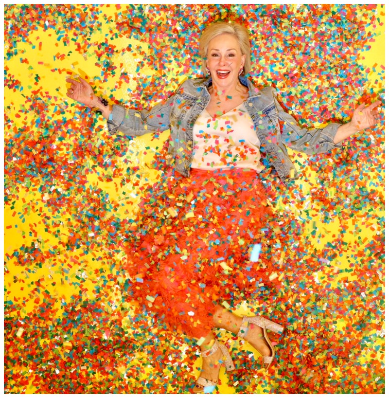 Sheree Frede of the SheShe Show wearing a bright coral midi length skirt, denim jacket over white camisole and rhinestone shoes laying on the floor covered in colorful confetti
