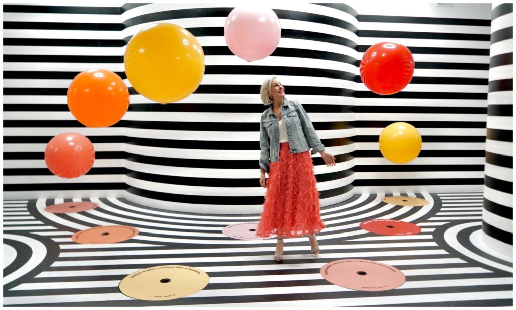 Sheree Frede of the SheShe Show wearing a bright coral midi length skirt, denim jacket over white camisole and rhinestone shoes staniding in a black and white strips room with large balloons floating around