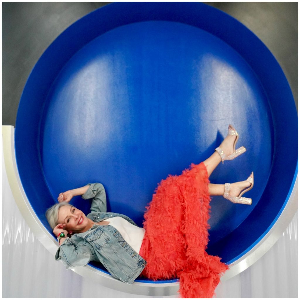 Sheree Frede of the SheShe Show wearing a bright coral midi length skirt, denim jacket over white camisole and rhinestone shoes laying on her back inside a large circle with blue background