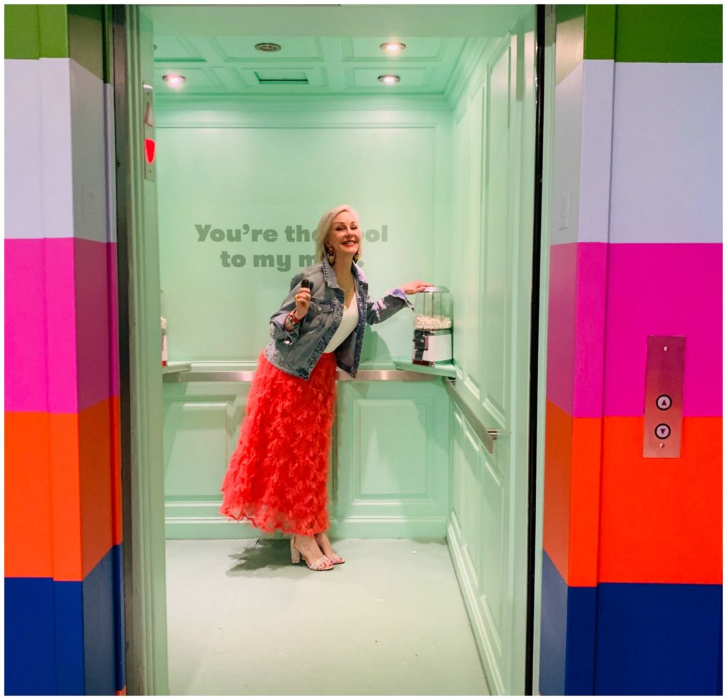 Sheree Frede of the SheShe Show wearing a bright coral midi length skirt, denim jacket over white camisole and rhinestone shoes standing inside a fun elevator