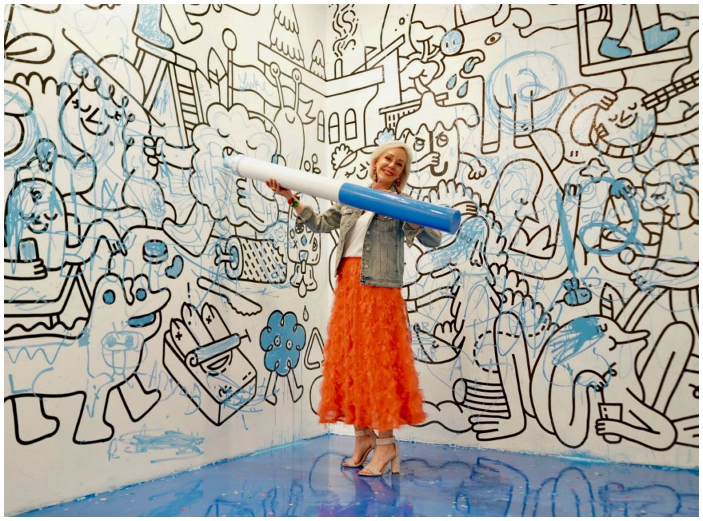 Sheree Frede of the SheShe Show wearing a bright coral midi length skirt, denim jacket over white camisole and rhinestone shoes standing in a drawing room holding a giant marker