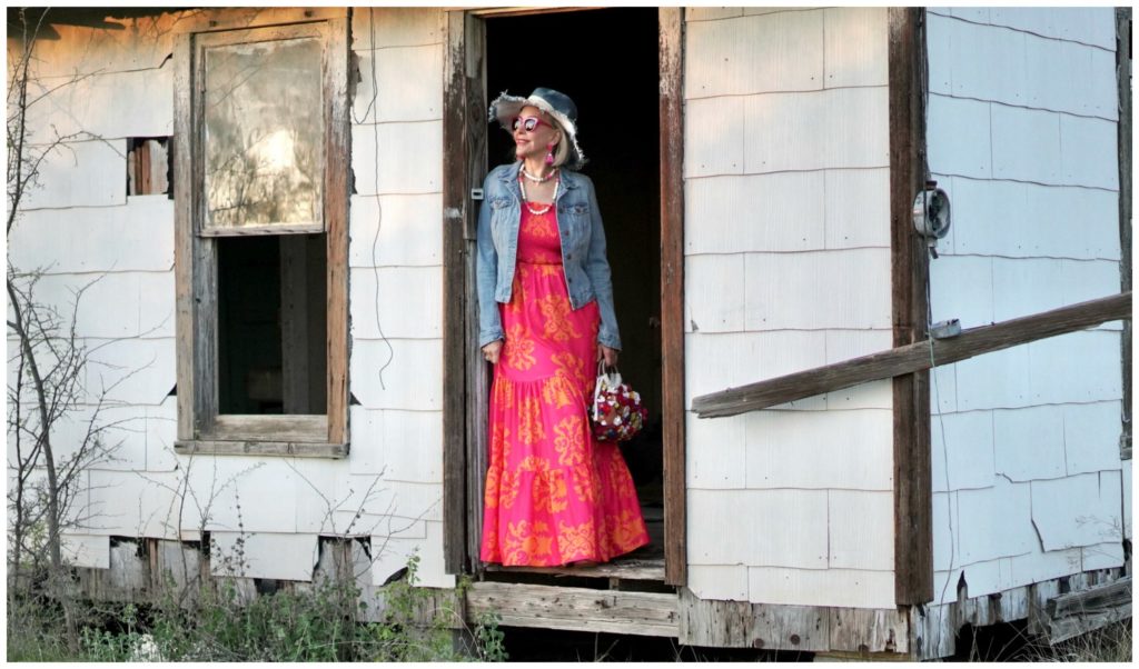 Sheree Frede of the SheShe Who standing in an abandoned house wearing an orange and pink maxi dress with denim jacket
