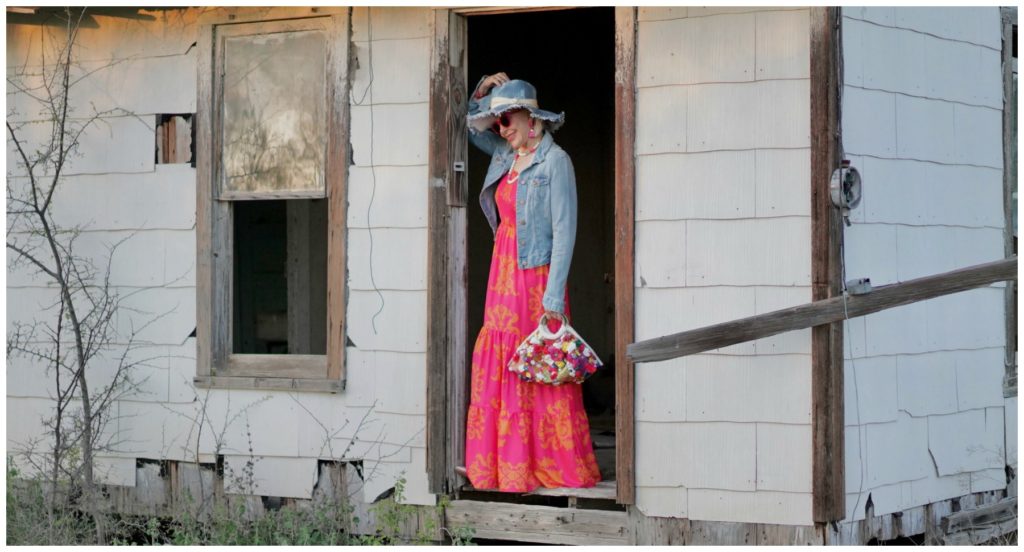 Sheree Frede of the SheShe Who standing in an abandoned house wearing an orange and pink maxi dress with denim jacket