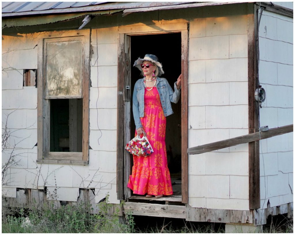 Sheree Frede of the SheShe Who standing in an abandoned house wearing an orange and pink maxi dress with denim jacket