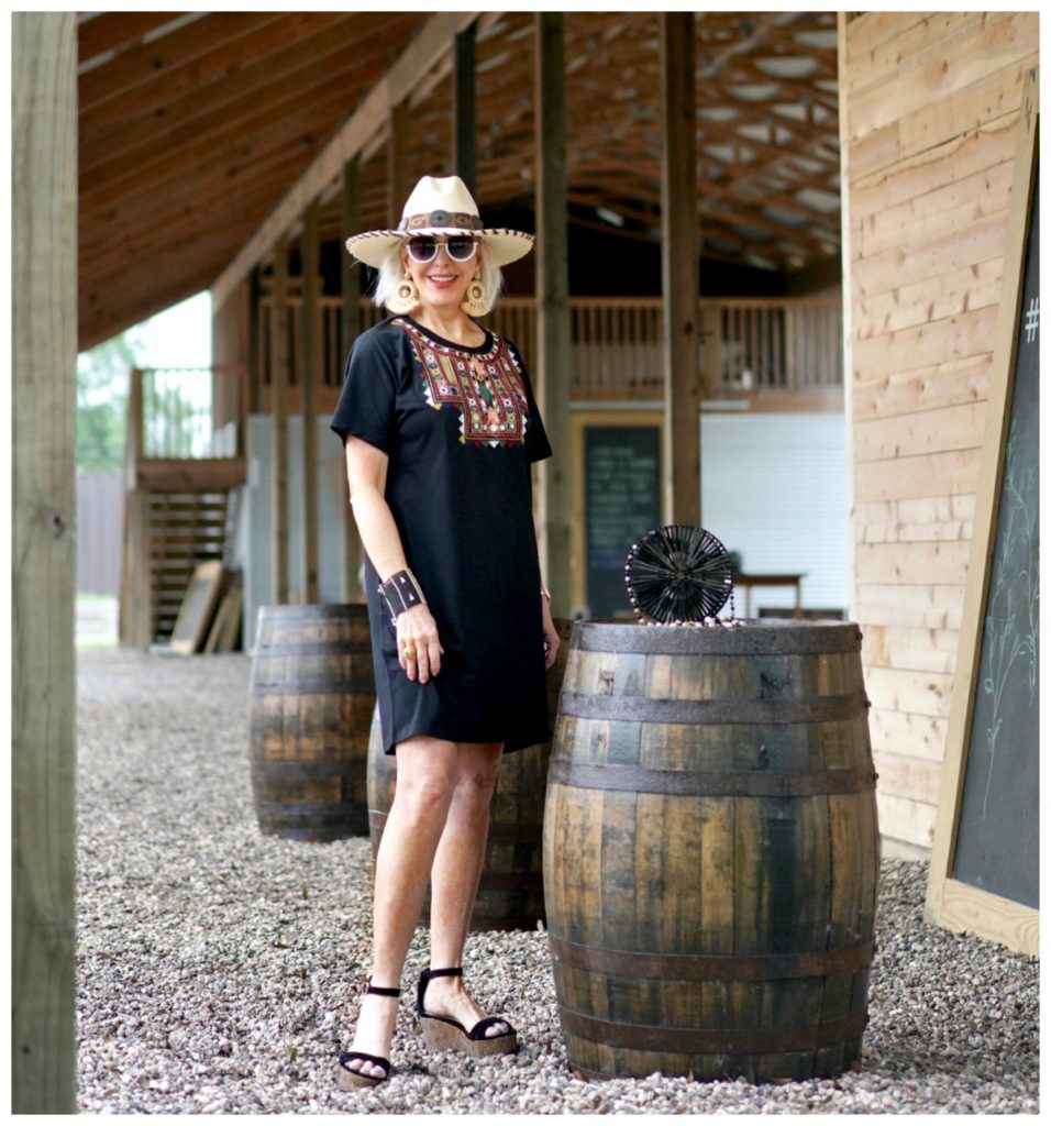 Sheree Frede of the SheShe Show standing under a shed by a barrel wearing a black shirt dress and hat