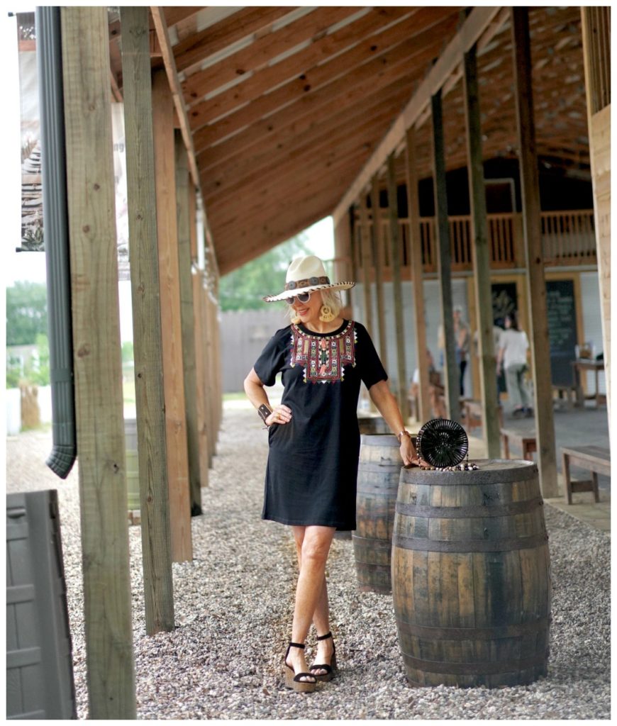 Sheree Frede of the SheShe Show standing under a shed by a barrel wearing a black shirt dress and hat