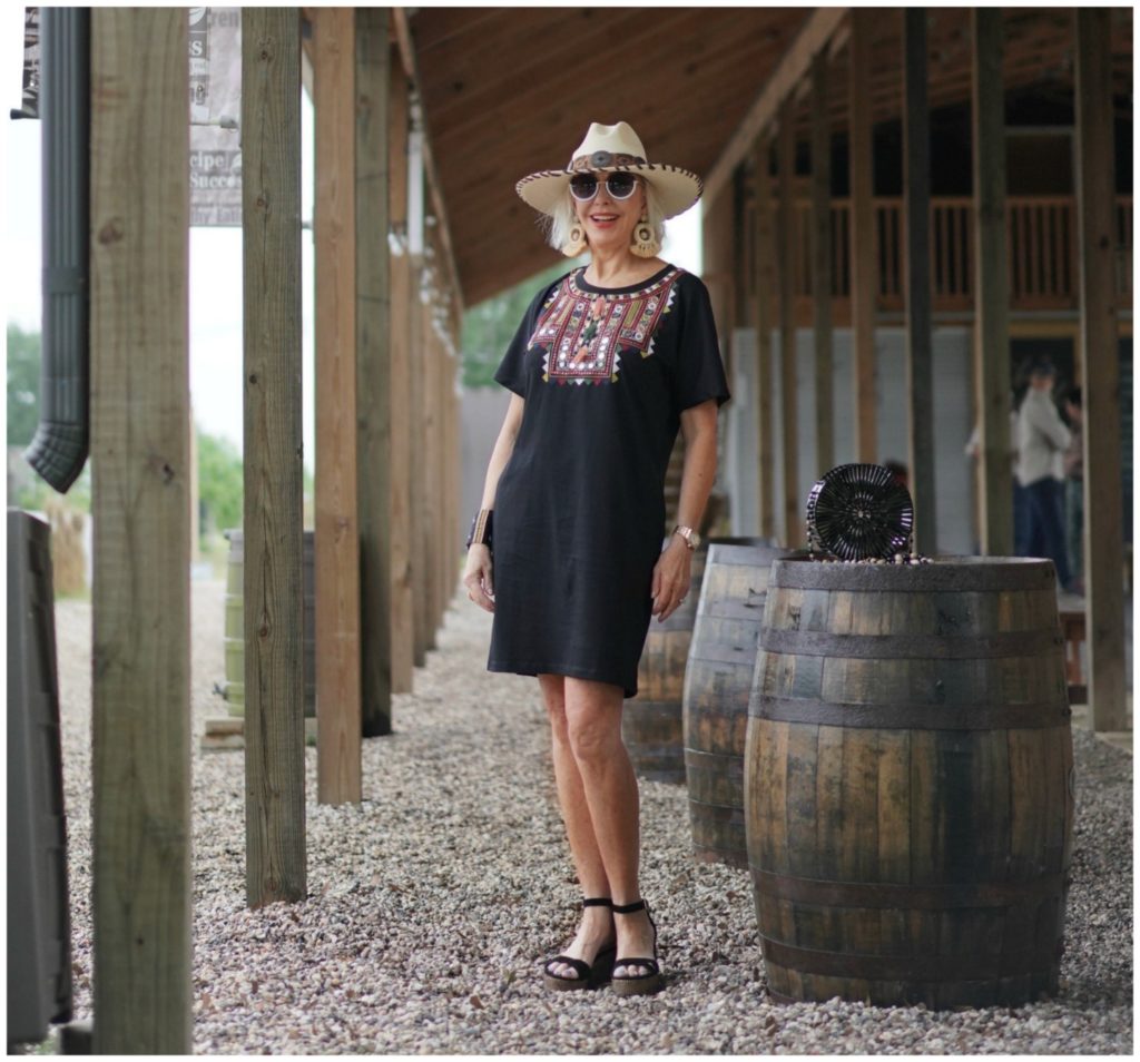 Sheree Frede of the SheShe Show standing under a shed by a barrel wearing a black shirt dress and hat