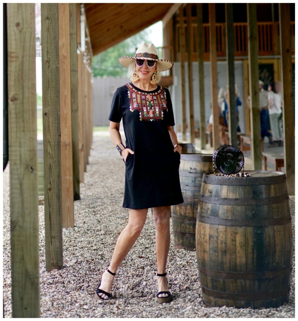 Sheree Frede of the SheShe Show standing under a shed by a barrel wearing a black shirt dress and hat