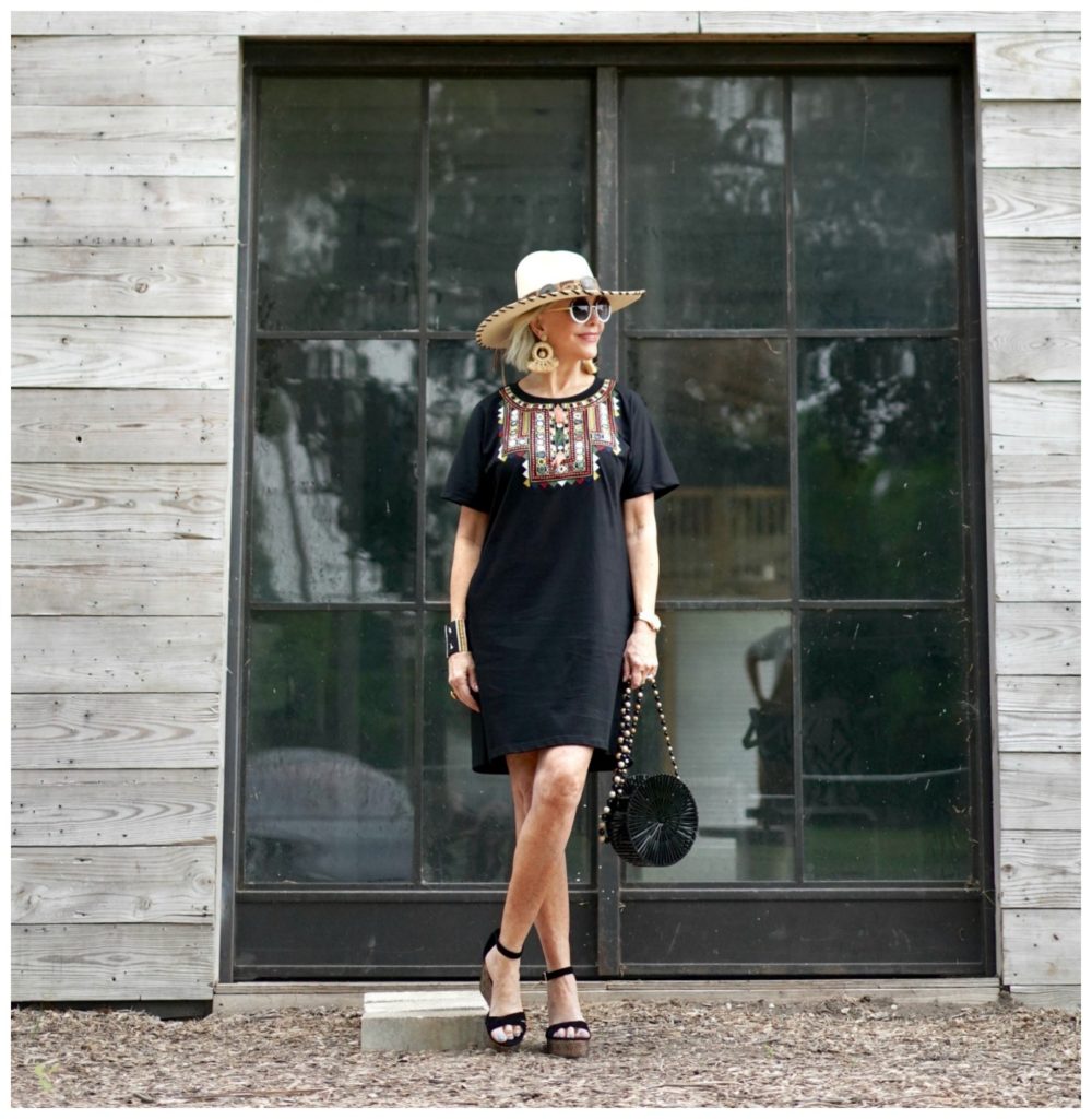 Sheree Frede of the SheShe Show standing under a shed by a barrel wearing a black shirt dress and hat