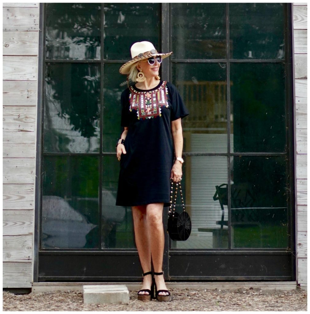 Sheree Frede of the SheShe Show standing under a shed by a barrel wearing a black shirt dress and hat