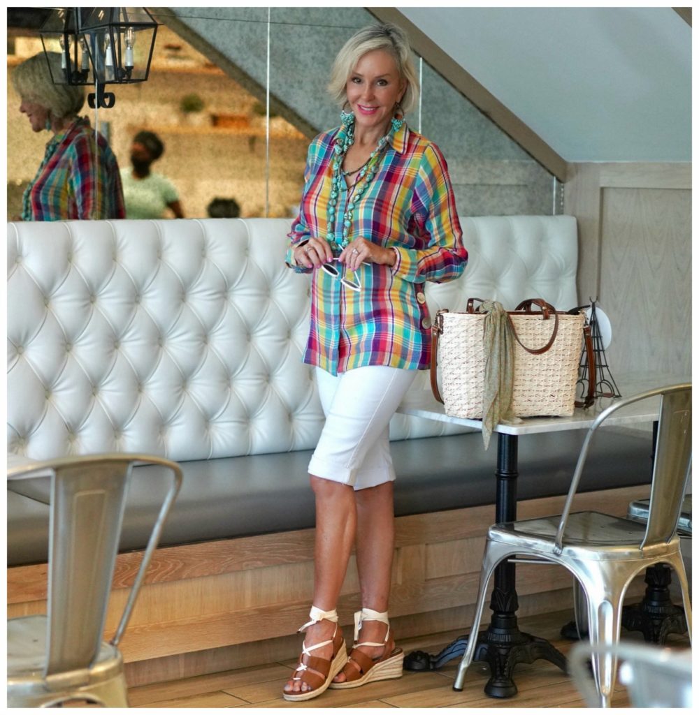 Sheree Frede of the SheShe SHow standing in a restaurant wearing a plaid shirt, white shorts, straw bag.