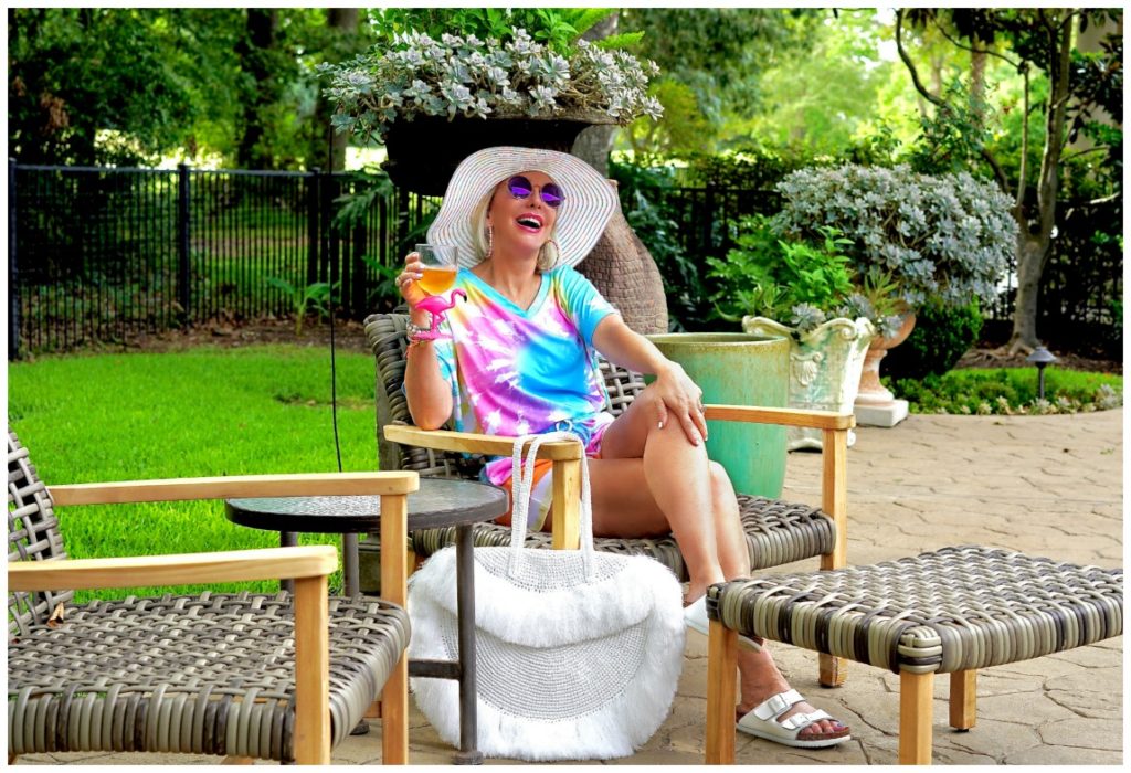 Sheree Frede of the SheShe Show out on patio wearing a 2 piece shorts and top tie dye set with large white fringy straw tote bag and big white hat