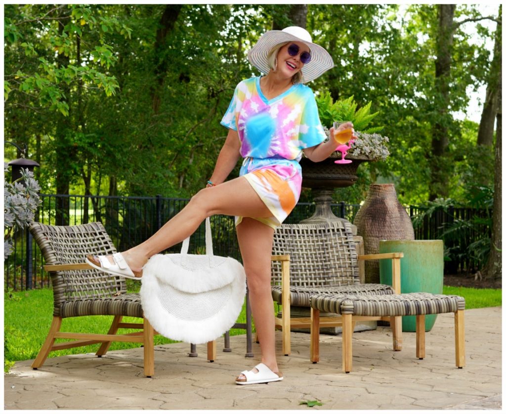 Sheree Frede of the SheShe Show out on patio wearing a 2 piece shorts and top tie dye set with large white fringy straw tote bag and big white hat