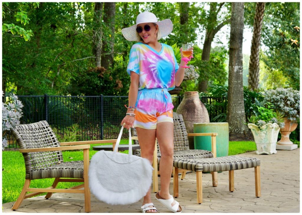 Sheree Frede of the SheShe Show out on patio wearing a 2 piece shorts and top tie dye set with large white fringy straw tote bag and big white hat