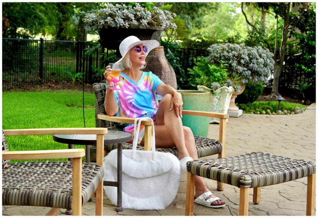 Sheree Frede of the SheShe Show out on patio wearing a 2 piece shorts and top tie dye set with large white fringy straw tote bag and big white hat