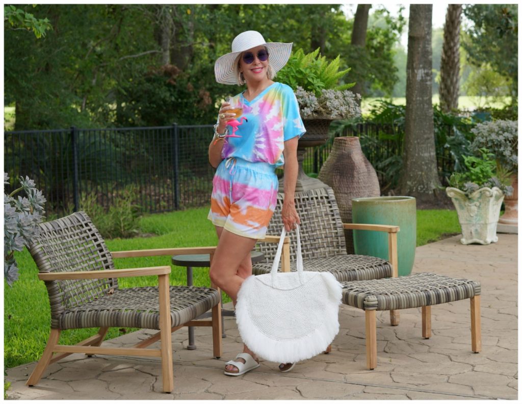 Sheree Frede of the SheShe Show out on patio wearing a 2 piece shorts and top tie dye set with large white fringy straw tote bag and big white hat