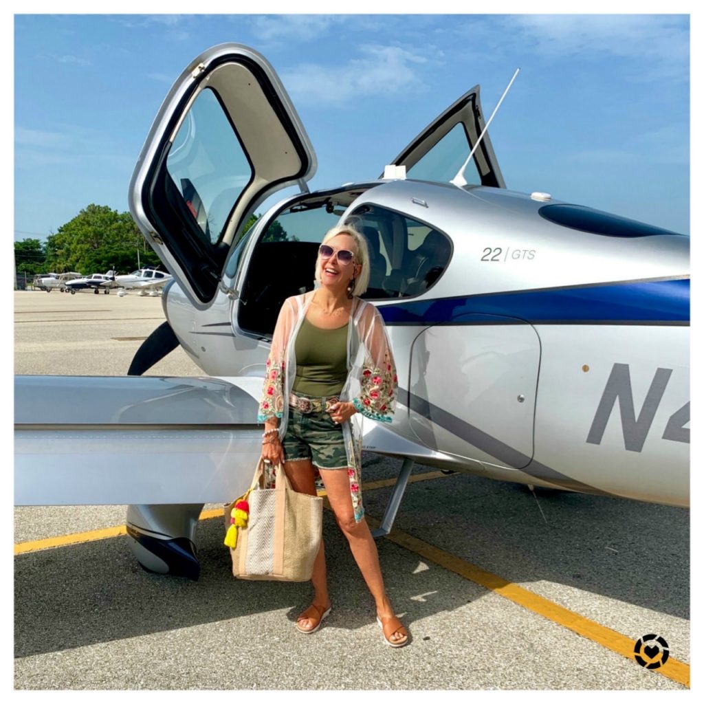 Sheree Frede of the SheShe Show standing in front of Cirrus aircraft wearing camo shorts and summer tote bag