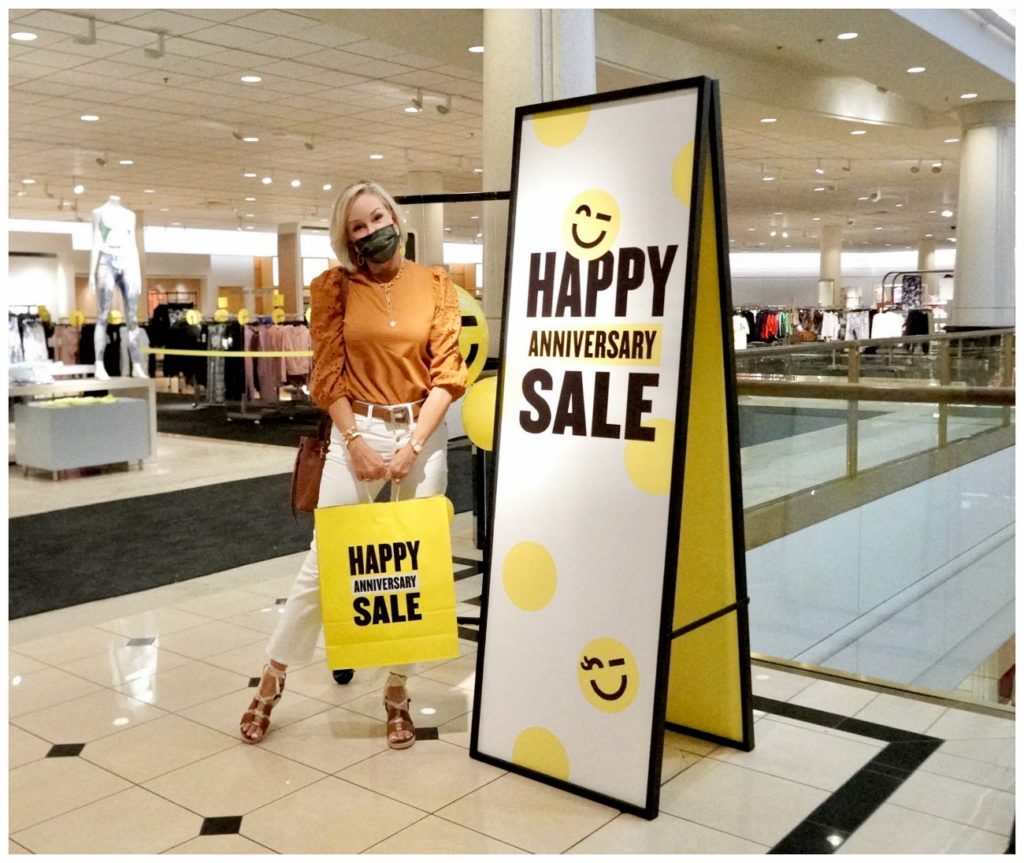 Sheree Frede of the SheShe Show standing by Nordstrom Anniversary Sale sign and holding yellow shopping bag