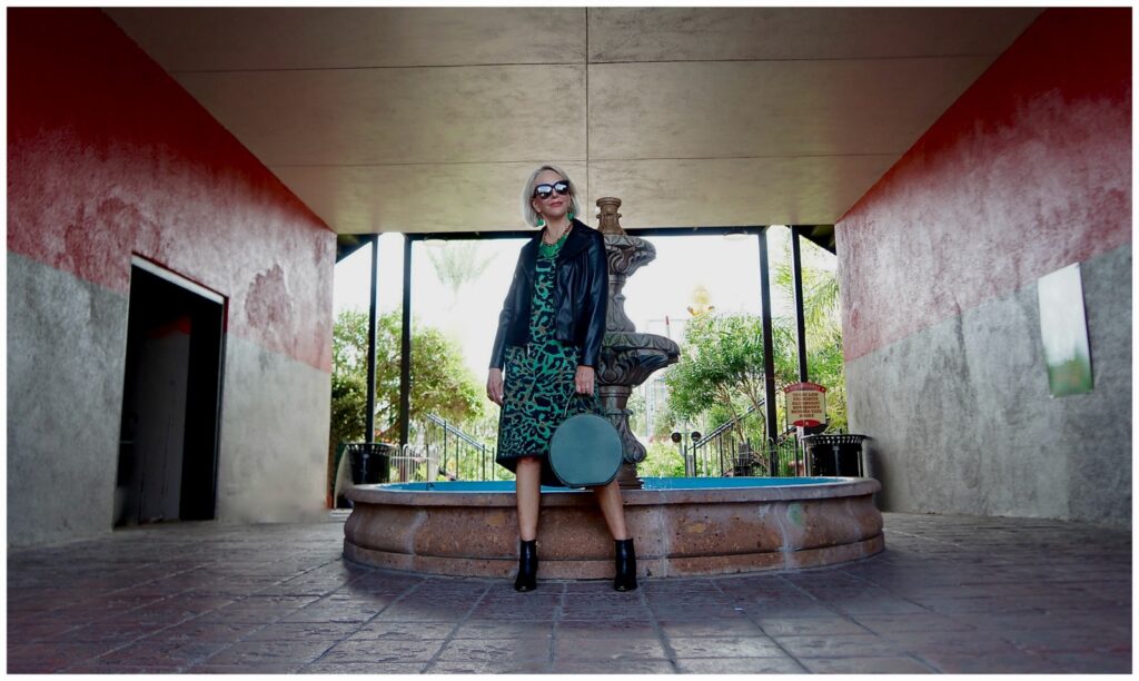 Sheree Frede of the SheShe Show walking in front of a a water fountain wearing a green leopard print dress with black leather jacket